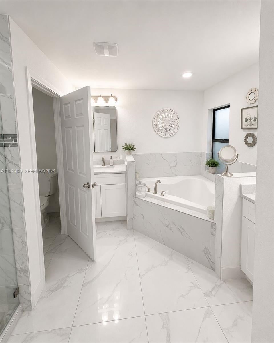 A white bathroom features a corner soaking tub, a vanity, and marble-look tile flooring — 1006 Rush Ave, Lehigh Acres FL 33972, LEHIGH ACRES, FL