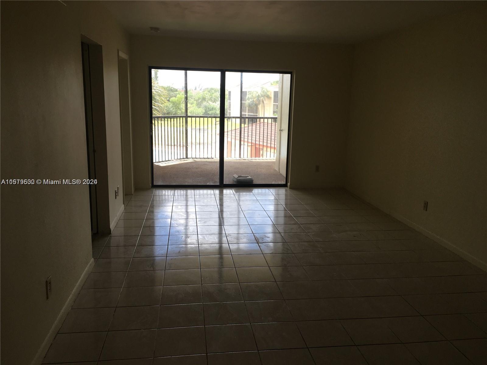 An empty room features light-colored floor tiles, beige walls, and a sliding glass door to a balcony — 3396 Foxcroft Rd # 308, Miramar FL 33025, MIRAMAR, FL