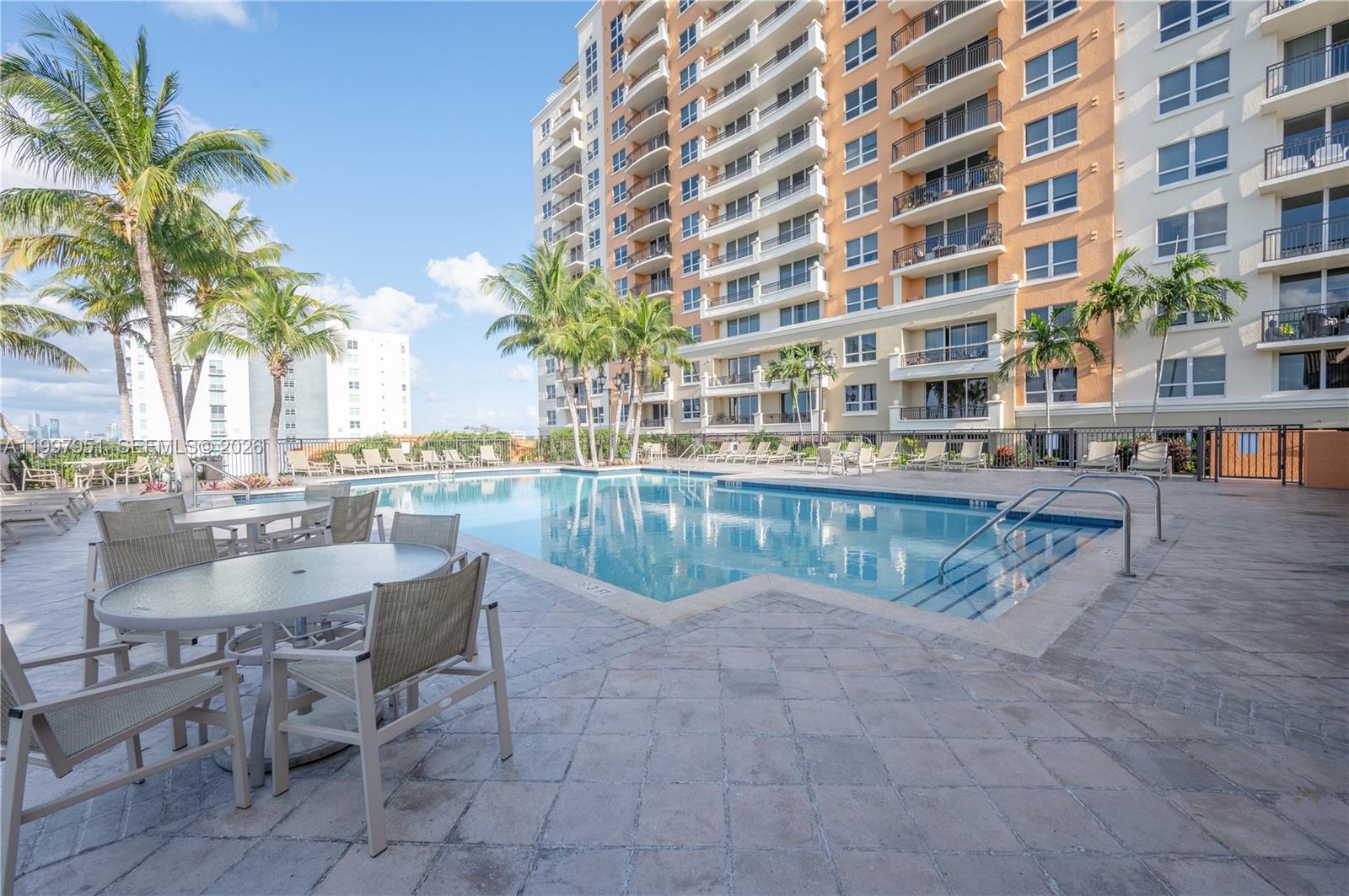 An outdoor swimming pool area with lounge chairs, tables, palm trees, and a tall building — 3232 SW 22nd Ter # 607, Miami FL 33145, MIAMI, FL