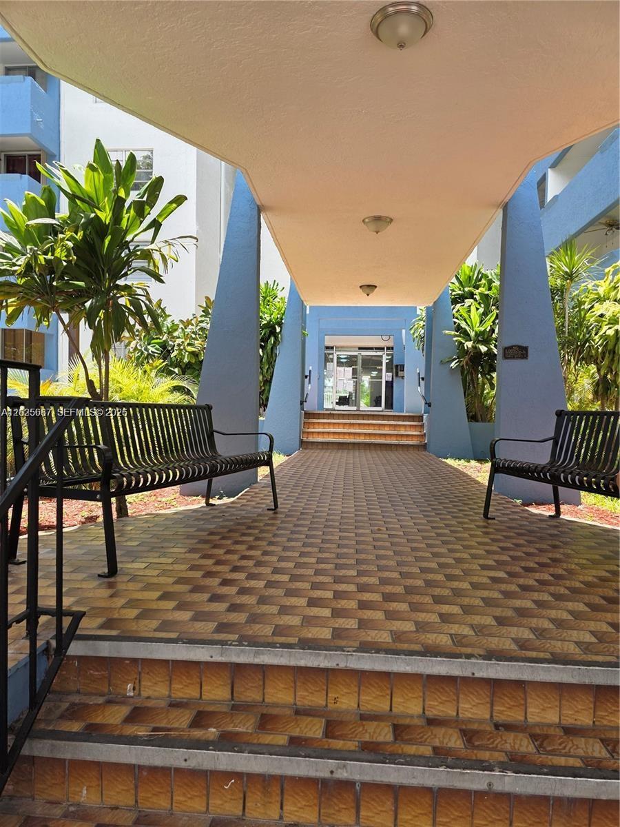A covered walkway with benches leads to the glass entrance of a blue apartment building — 5050 NW 7th St # 610, Miami FL 33126, MIAMI, FL