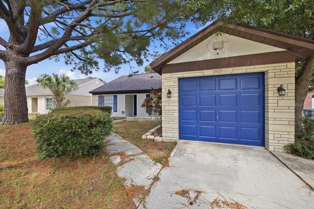 A single-story house with a blue garage door, stone facade, and a large pine tree — 1901 CASTLE BAY COURT, OLDSMAR, FL