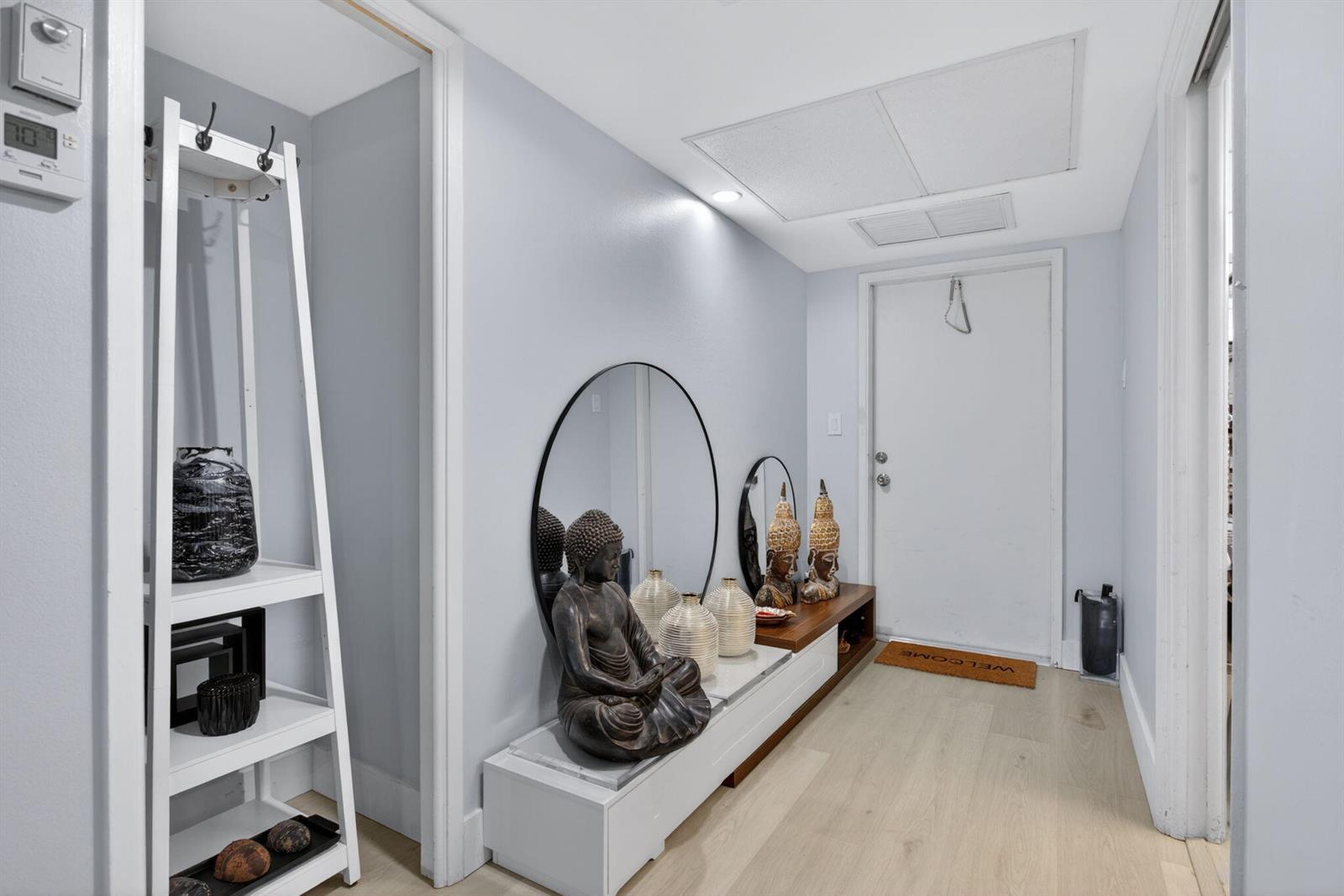 A modern entryway features light wood flooring, a white console table, mirrors, and Buddha statues — 500 Oaks Lane # 111, Pompano Beach FL 33069, POMPANO BEACH, FL