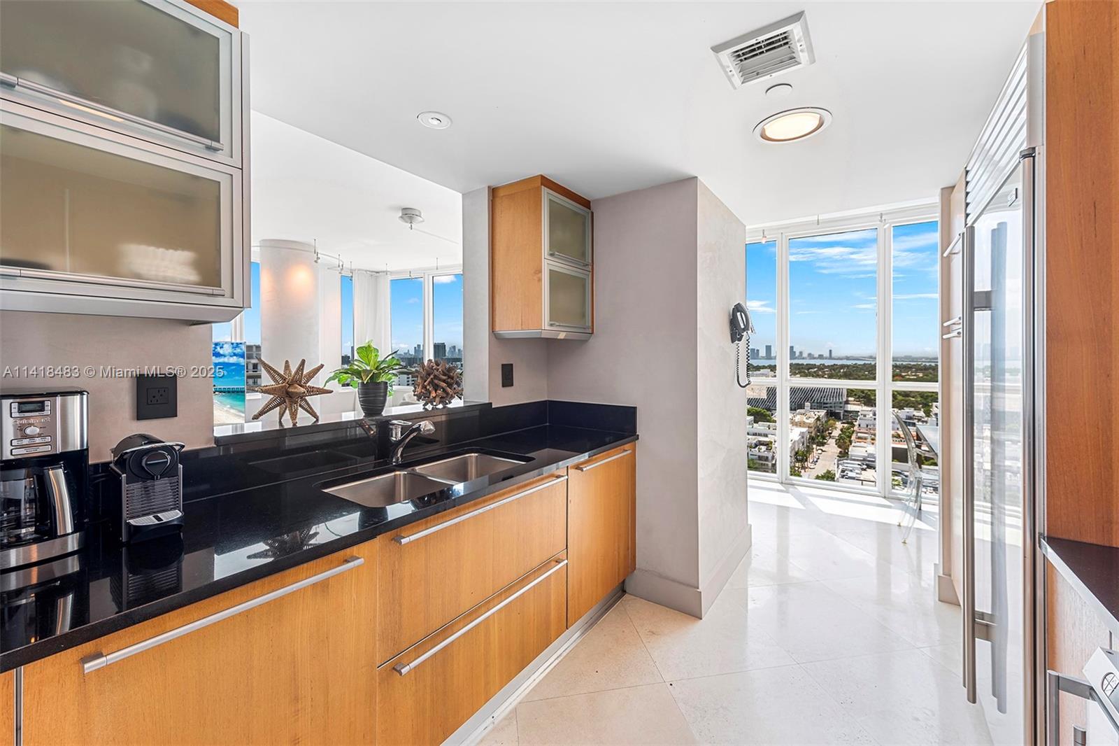 A kitchen features wood cabinets, black countertops, stainless steel appliances, and floor-to-ceiling windows with city views — 101 20th St # 2102, Miami Beach FL 33139, MIAMI BEACH, FL