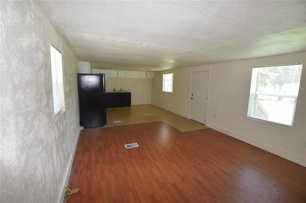 An open-concept room features wood-look flooring, a kitchen area with dark cabinets, and white walls — 5101 N KALIGA DRIVE, ST CLOUD, FL