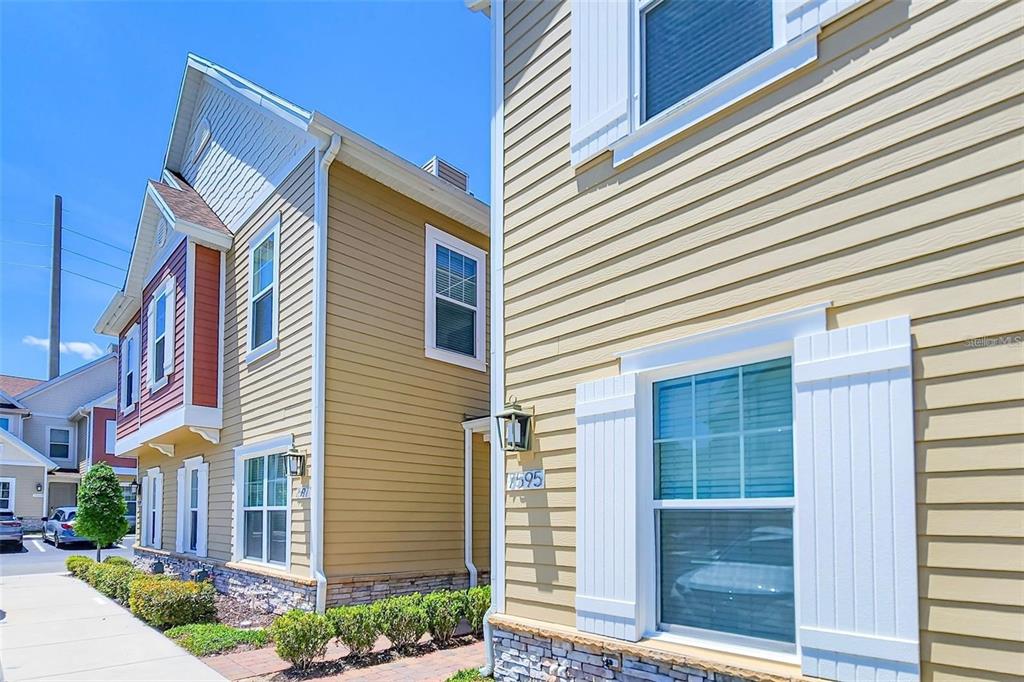 A low-angle exterior view of tan-sided townhomes with white shutters and stone accents — 7595 SUNNY DREAMS LANE, KISSIMMEE, FL
