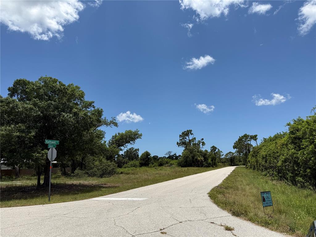 An empty, paved road curves through a grassy, wooded lot under a bright blue sky — 12526 RATHWELL AVENUE, PORT CHARLOTTE, FL