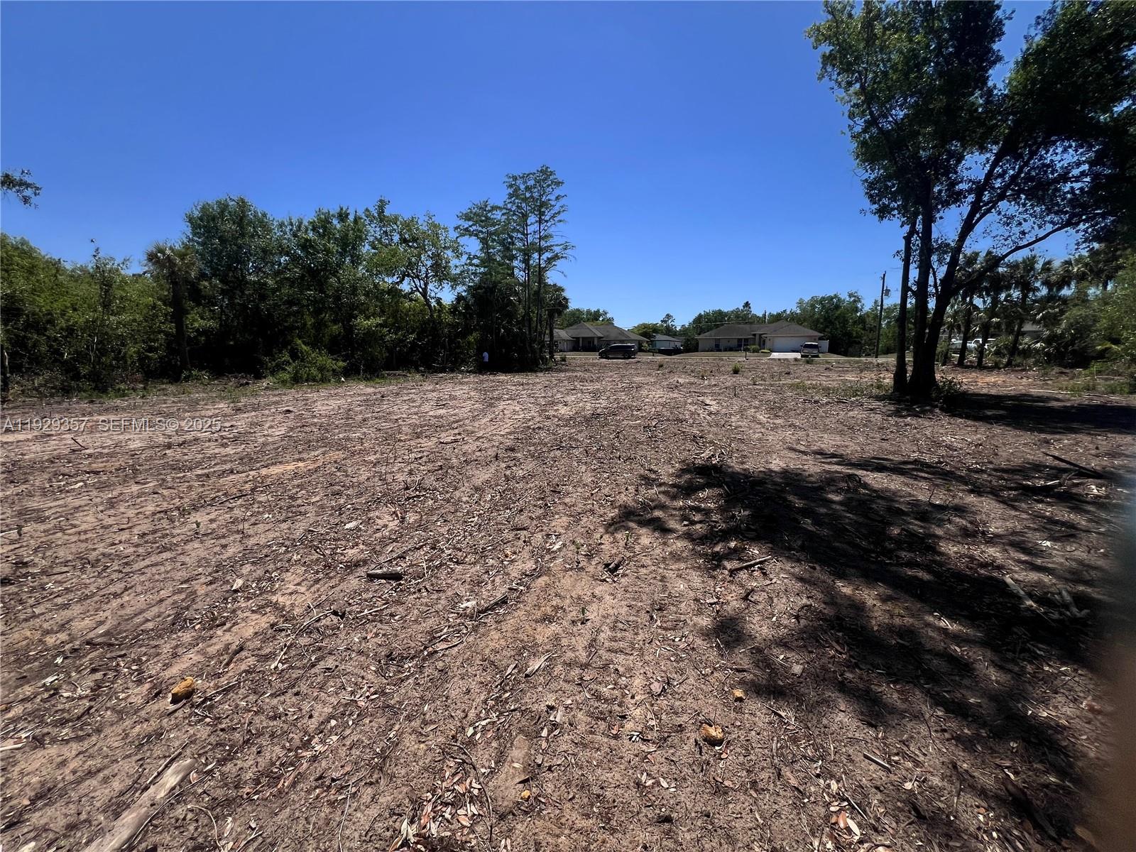 A cleared, dirt-covered vacant lot is surrounded by trees under a clear blue sky — 3414 W 32nd, Lehigh Acres FL 33971, LEHIGH ACRES, FL
