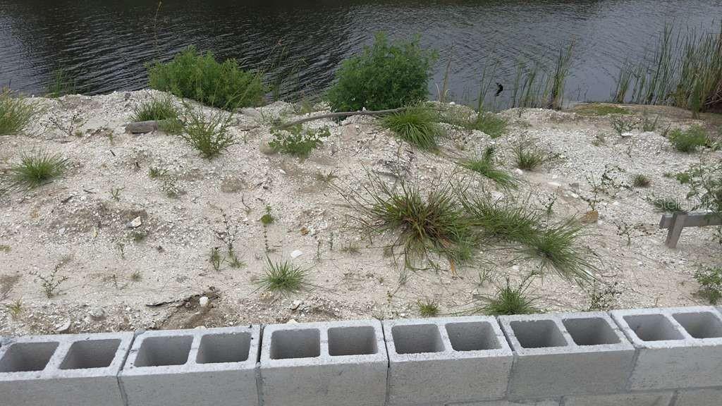 A low concrete block wall sits in front of a sandy, sparsely vegetated waterfront lot — 15408 LAKELAND CIRCLE, PORT CHARLOTTE, FL