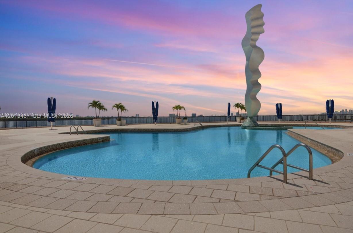 An outdoor swimming pool features a tall abstract sculpture against a sunset sky — 1800 NE 114th St # 601, Miami FL 33181, MIAMI, FL