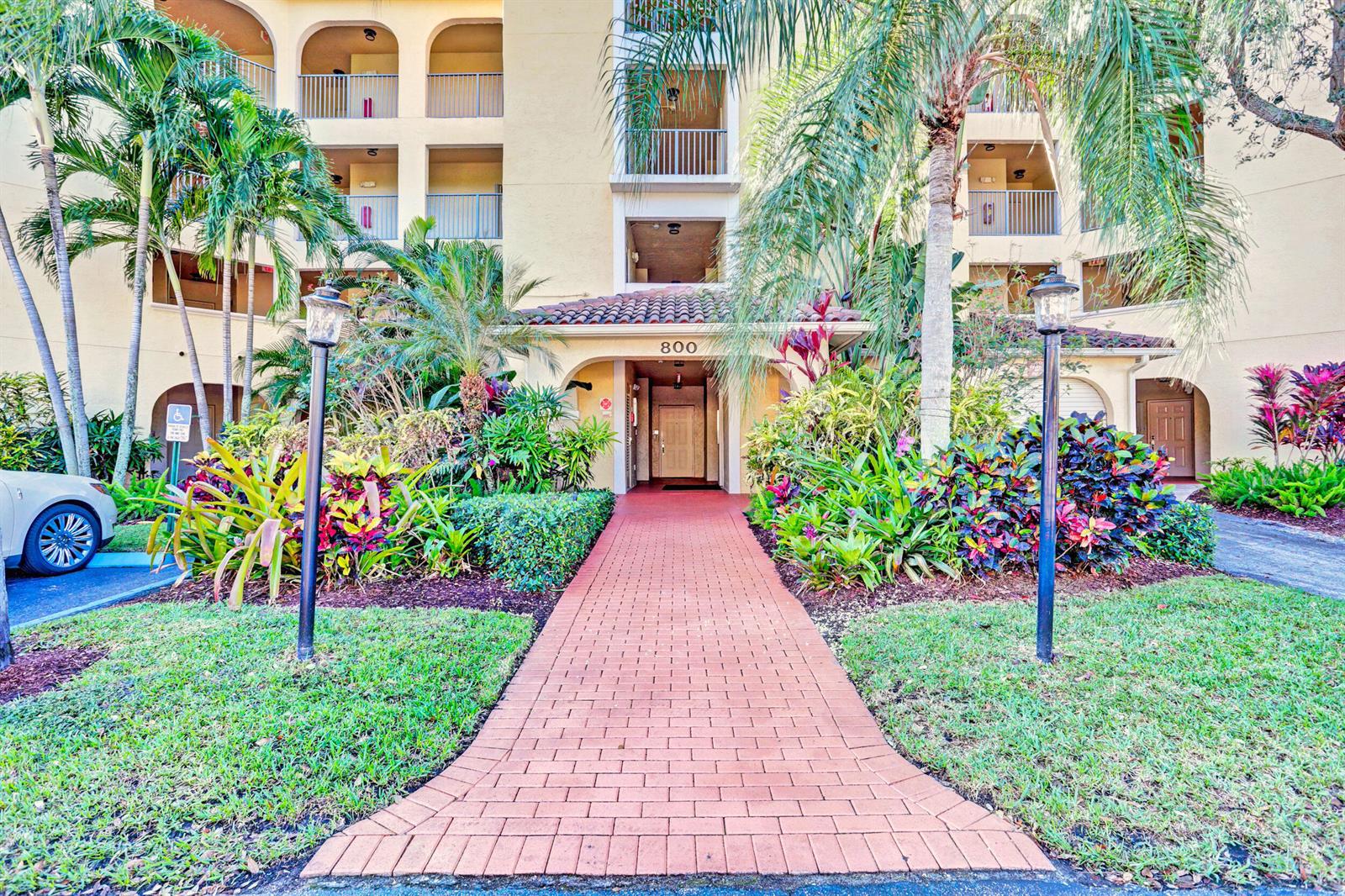 A brick walkway leads to the entrance of a multi-story apartment building with tropical landscaping — 800 Uno Lago Drive # 305, Juno Beach FL 33408, JUNO BEACH, FL