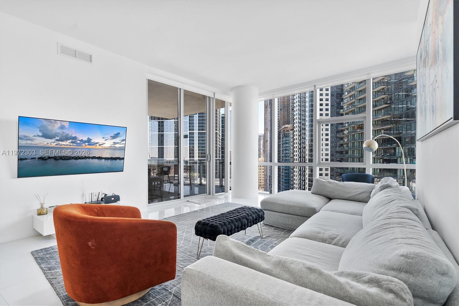 A modern living room features a grey sectional, orange armchair, and floor-to-ceiling city views — 851 NE 1st Ave # 1712, Miami FL 33132, MIAMI, FL