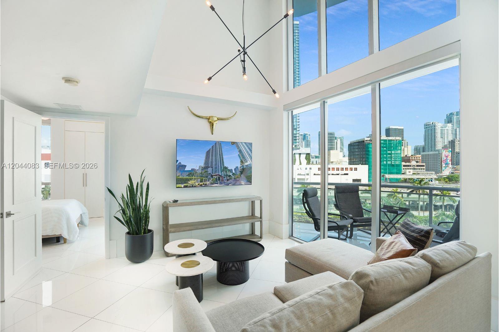 A modern living room with high ceilings, white tile floors, and a balcony overlooking city buildings — 350 S Miami Ave # 713, Miami FL 33130, MIAMI, FL