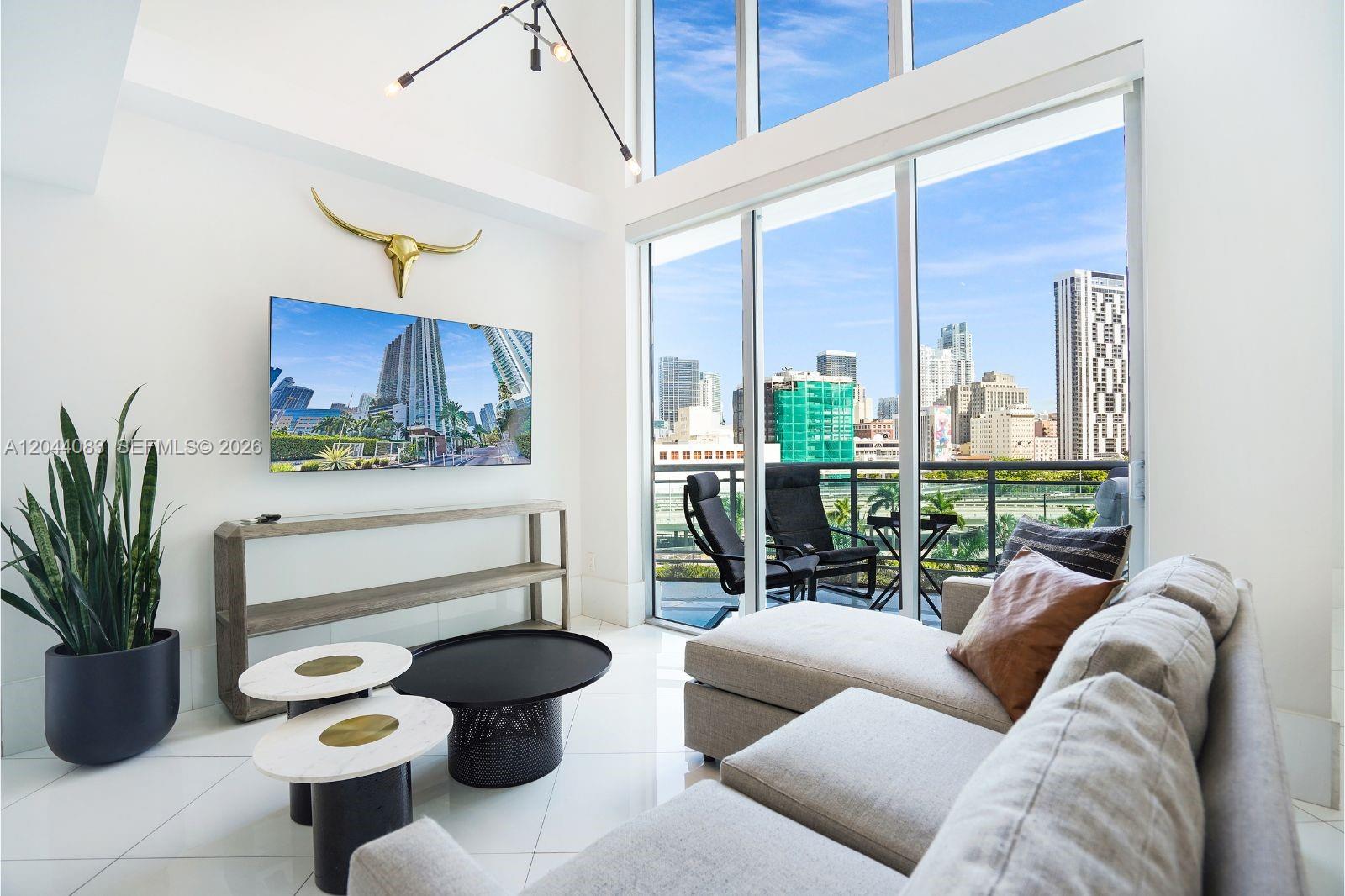 A modern living room features a sectional sofa, television, and floor-to-ceiling windows overlooking a city — 350 S Miami Ave # 713, Miami FL 33130, MIAMI, FL