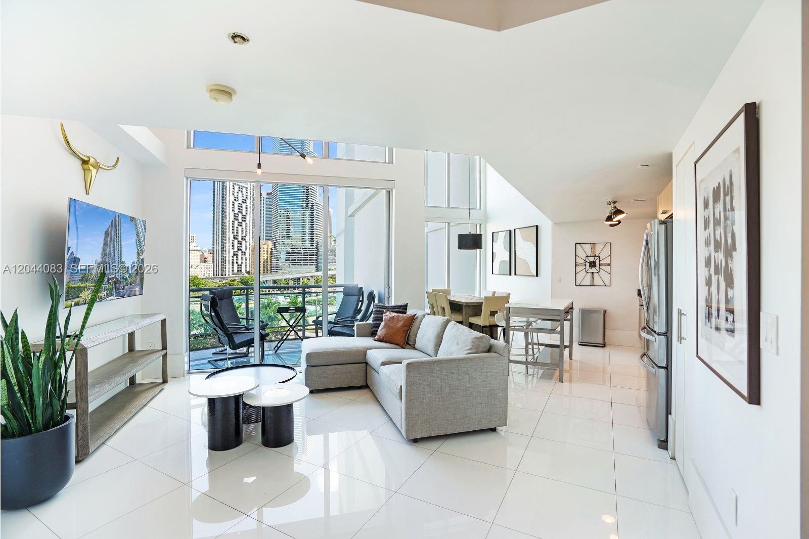 A modern living room features white tile floors, a gray sectional, and floor-to-ceiling city views — 350 S Miami Ave # 713, Miami FL 33130, MIAMI, FL