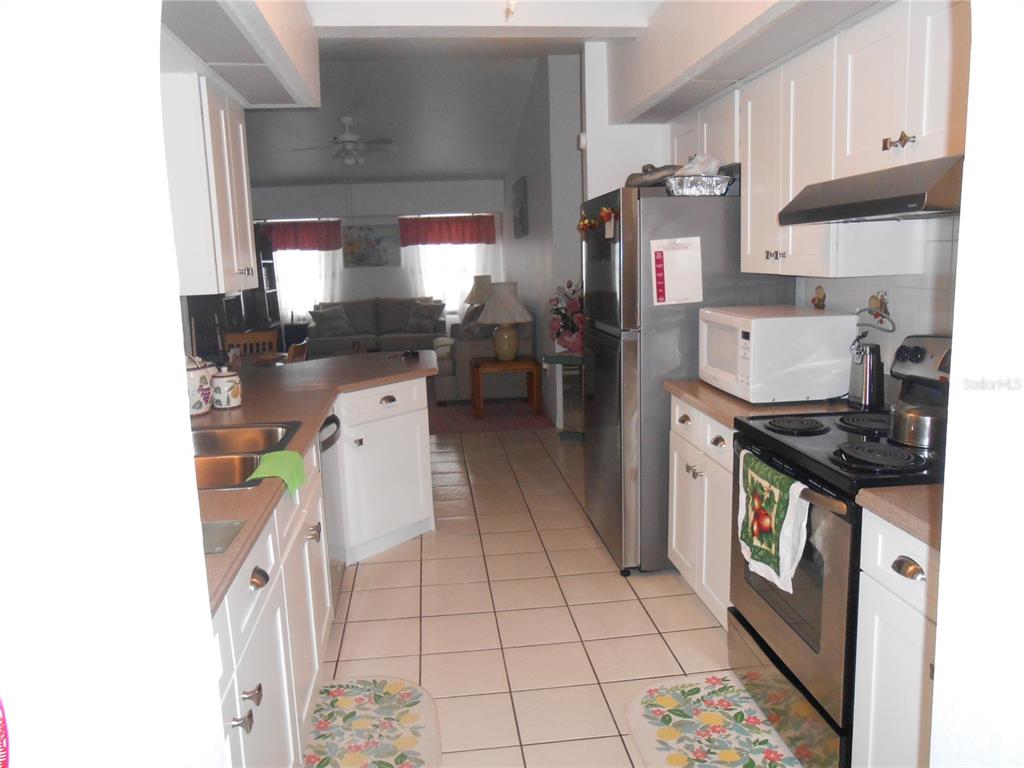 A galley kitchen with white cabinets, stainless steel appliances, and tile flooring leads to a living room — 21239 GAYLORD AVENUE, PORT CHARLOTTE, FL