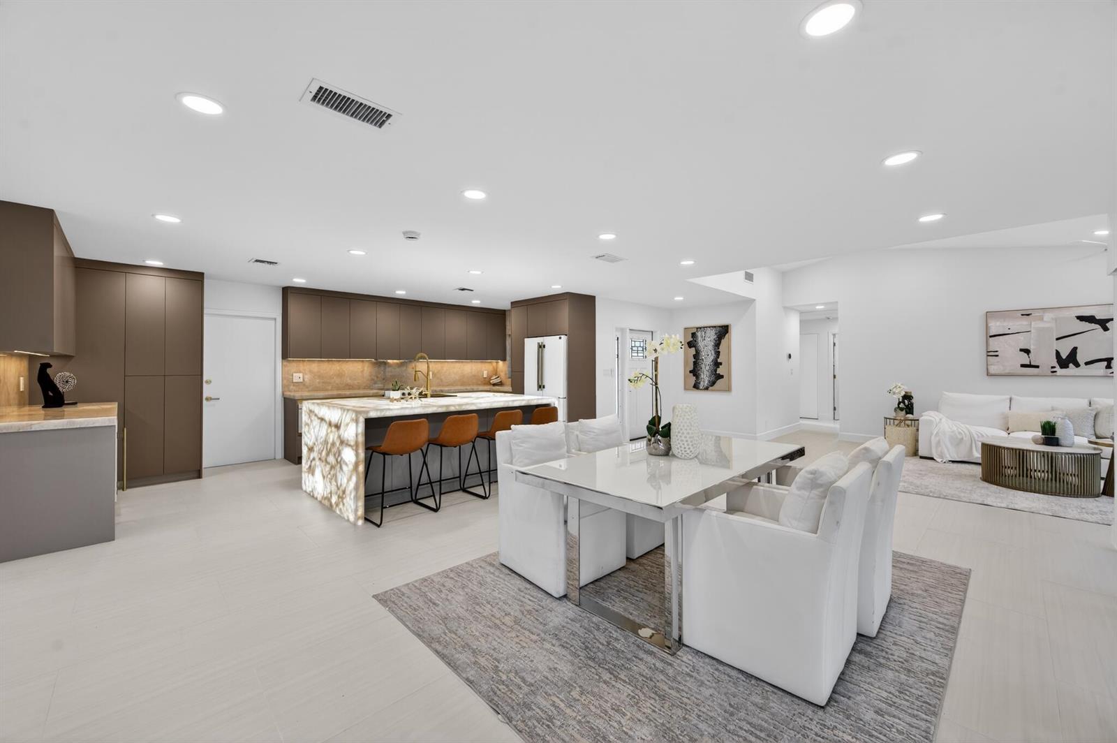 A modern open-concept kitchen and dining area features brown cabinetry, a backlit stone island, and white furniture — 3088 NW 26th Avenue, Boca Raton FL 33434, BOCA RATON, FL
