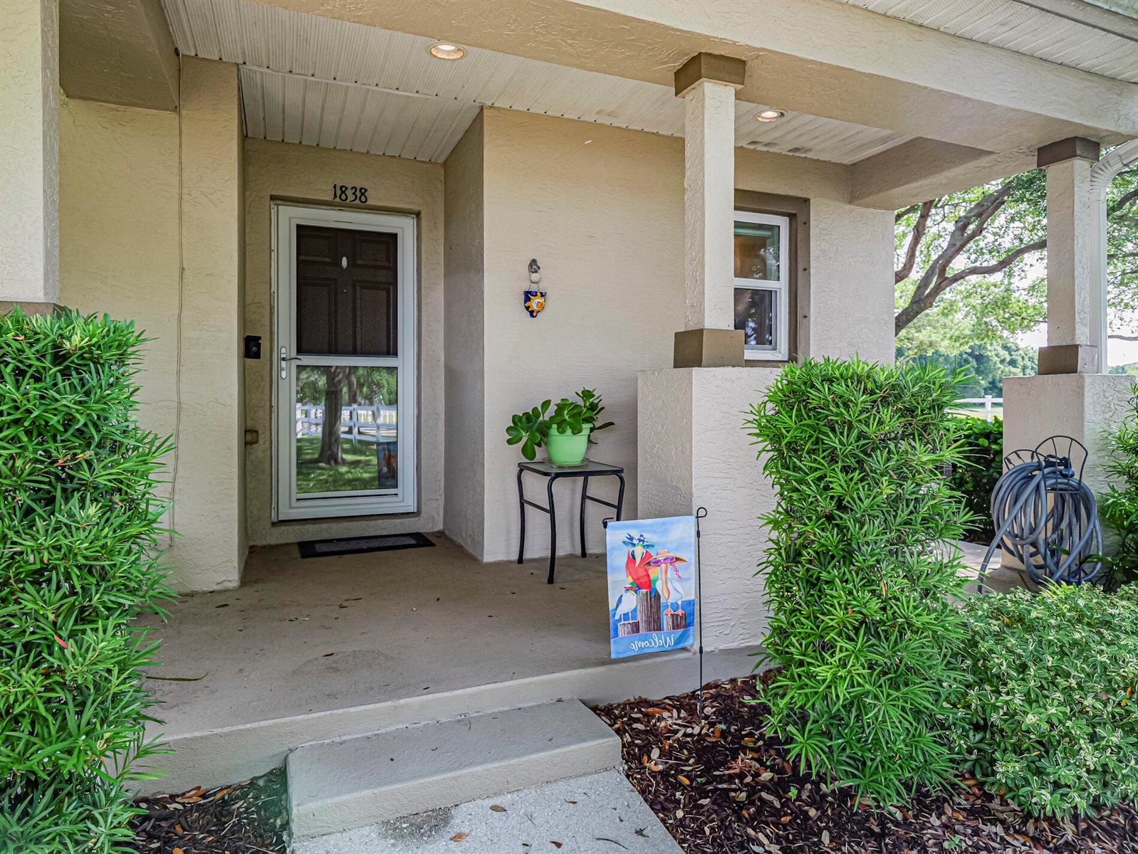 A beige house exterior features a front porch, a glass storm door, and manicured landscaping — 1838 Pointe West Way, Vero Beach FL 32966, VERO BEACH, FL