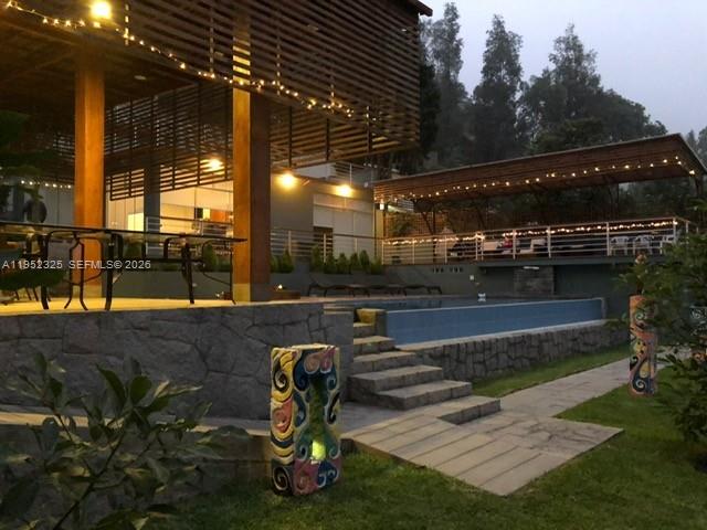 A modern house features a swimming pool, stone retaining walls, and string lights at dusk — 130 Av. Malecon, California, Chosica, Lima - Peru, FL, FL