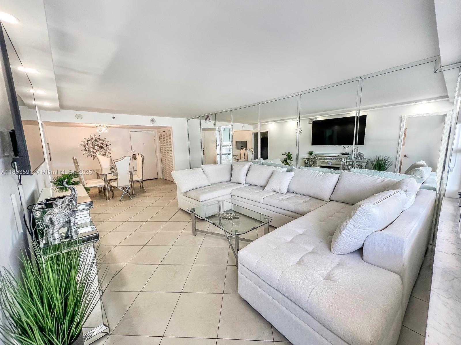 A living room features a large white sectional sofa, mirrored walls, and tile flooring — 5225 Collins Ave # 517, Miami Beach FL 33140, MIAMI BEACH, FL