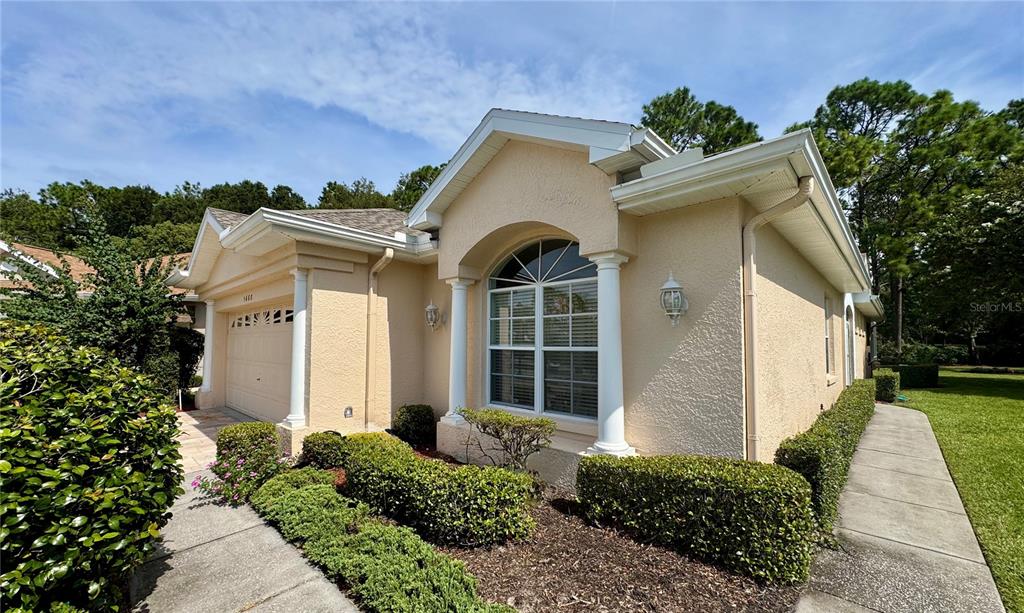 A tan stucco house features a front-facing garage, a large arched window, and landscaped hedges — 5688 LEGEND HILLS LANE, SPRING HILL, FL