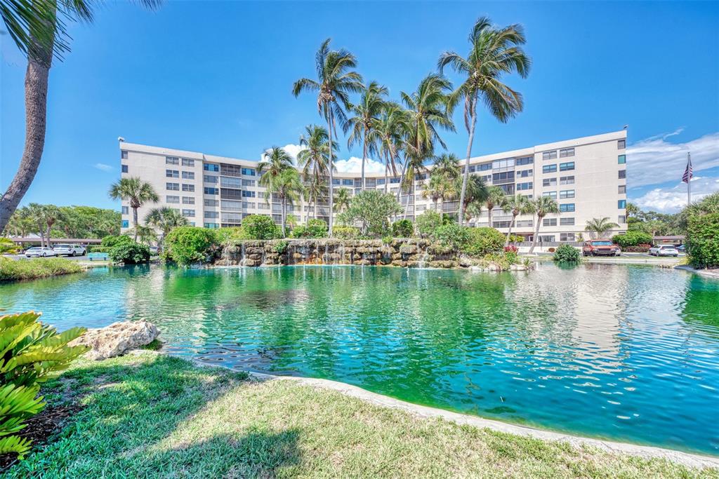 A multi-story apartment building sits behind a pond with a rock waterfall and palm trees — 5855 MIDNIGHT PASS ROAD UNIT 426, SARASOTA, FL
