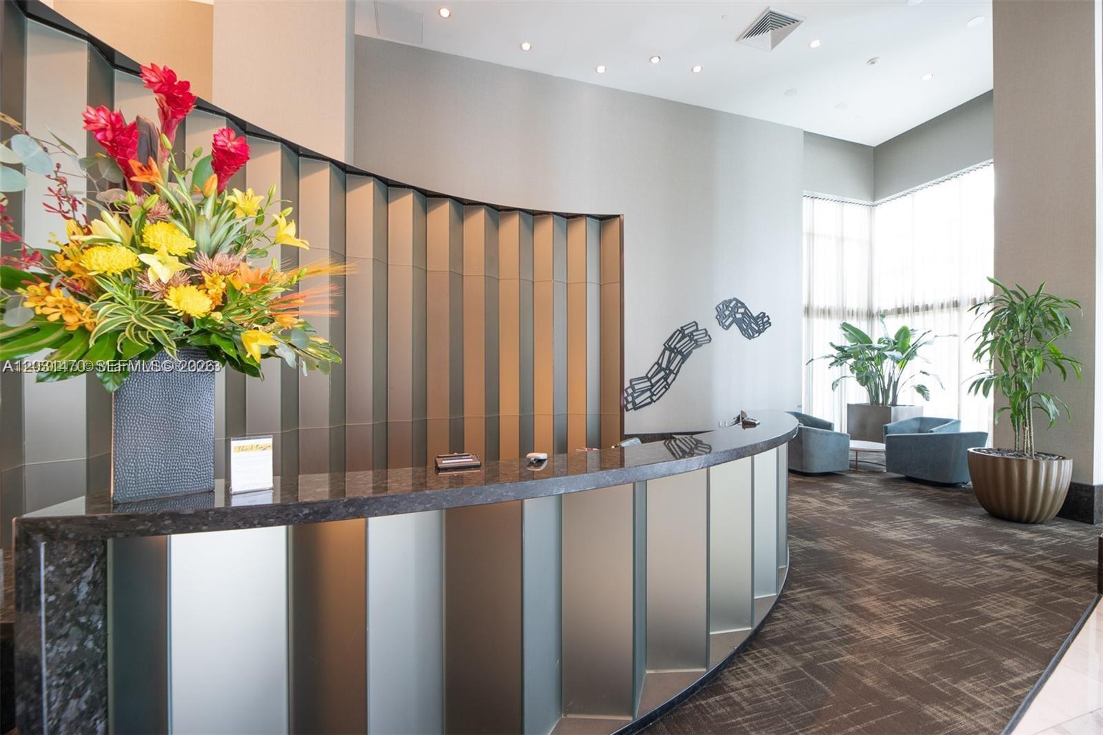 A modern reception area features a curved desk with metallic panels and a large floral arrangement — MIAMI, FL