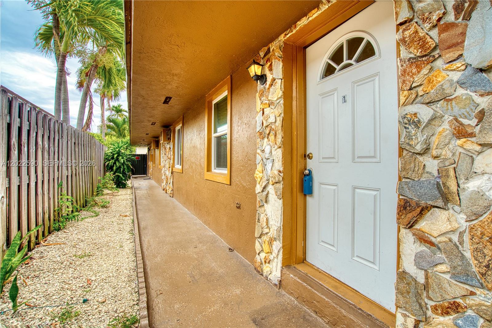 An exterior view shows a walkway along a tan building with stone accents and palm trees — 129 SE 4th Ave # 1, Hallandale Beach FL 33009, HALLANDALE BEACH, FL