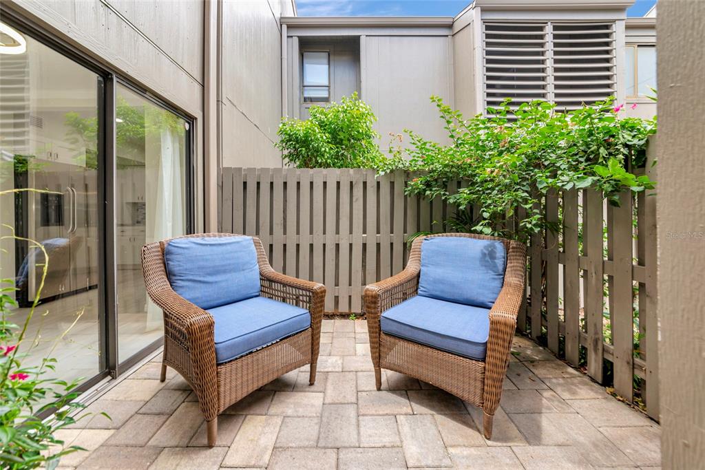 Two wicker armchairs with blue cushions sit on a paved patio enclosed by a fence — 108 WOODLAND PLACE UNIT 108, OSPREY, FL