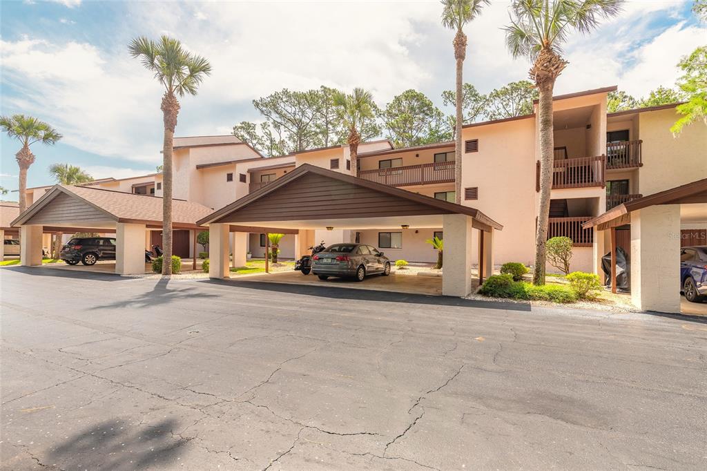 An exterior view of a multi-story apartment building with covered parking spaces and palm trees — 275 INDIGO DRIVE UNIT 204, DAYTONA BEACH, FL