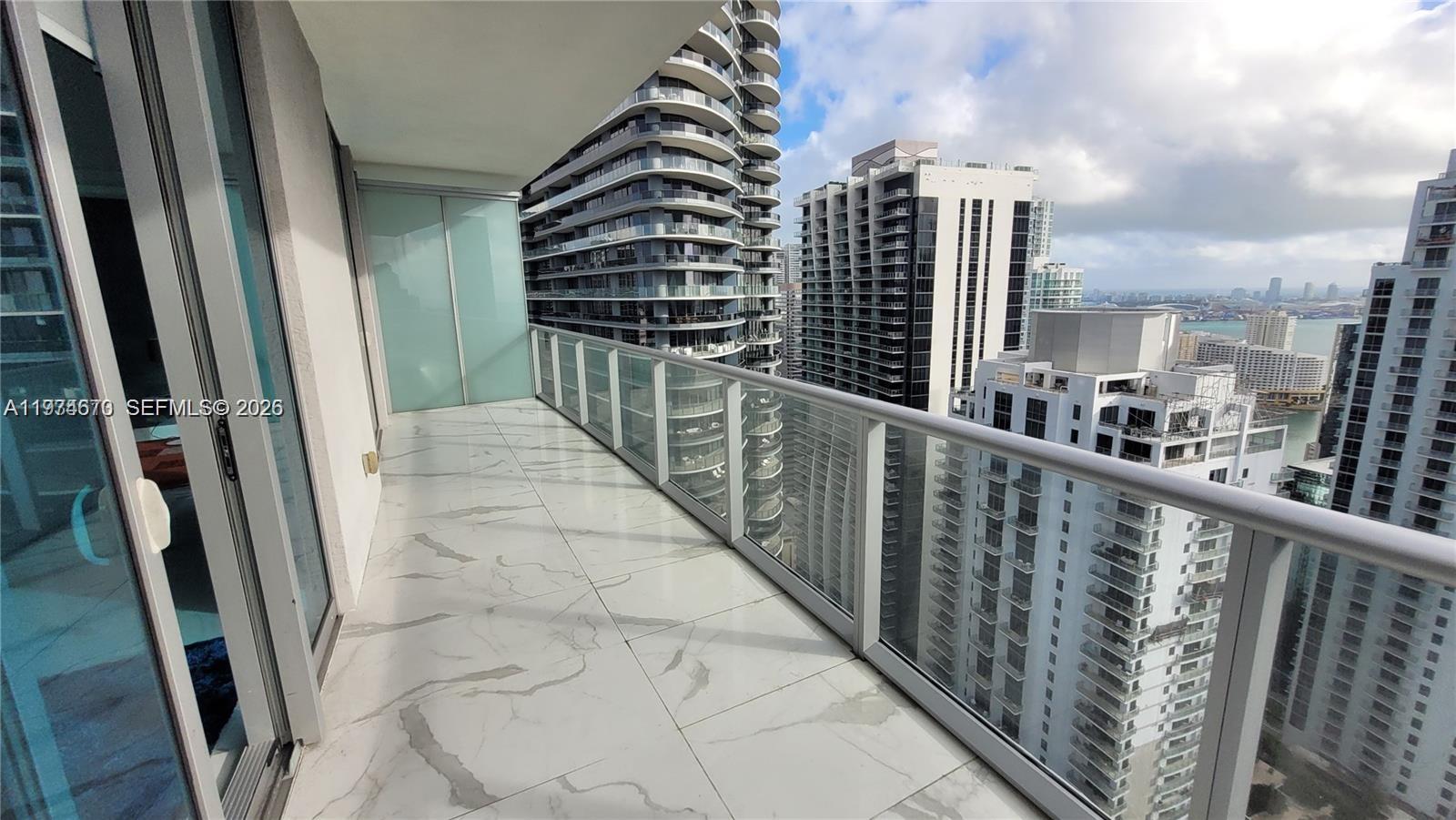 A high-rise balcony features white marble-look tile flooring and a glass railing overlooking city buildings — 1100 S Miami Ave # 4201, Miami FL 33130, MIAMI, FL