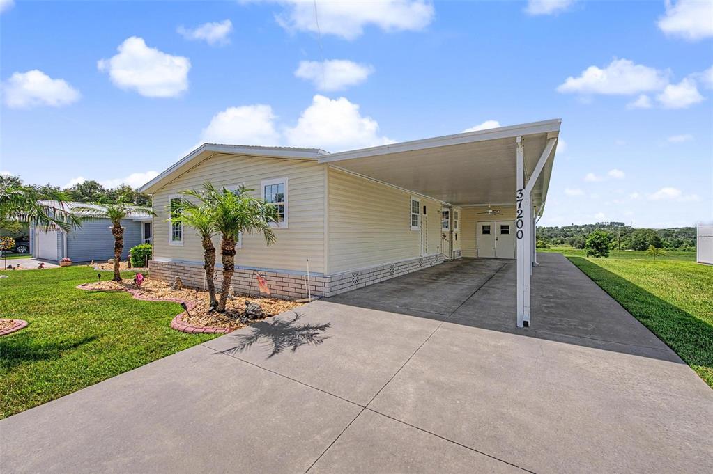 A light yellow manufactured home features a concrete driveway, a carport, and a small garden — 37200 NEUKOM AVENUE, ZEPHYRHILLS, FL