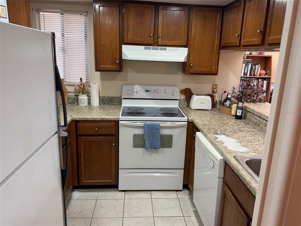 A kitchen features wood cabinets, white appliances, a speckled countertop, and a tiled floor — 1051 S HIGHLAND STREET UNIT 3D, MOUNT DORA, FL