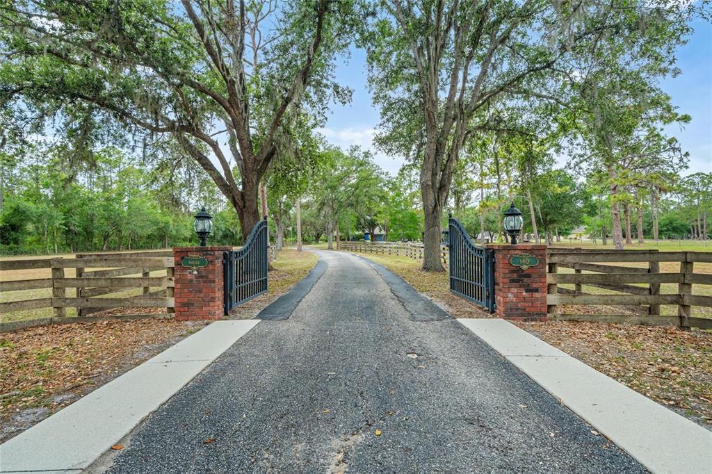 A paved driveway leads through an open brick-pillared gate into a wooded residential property — 140 COWPEN LANE, SARASOTA, FL