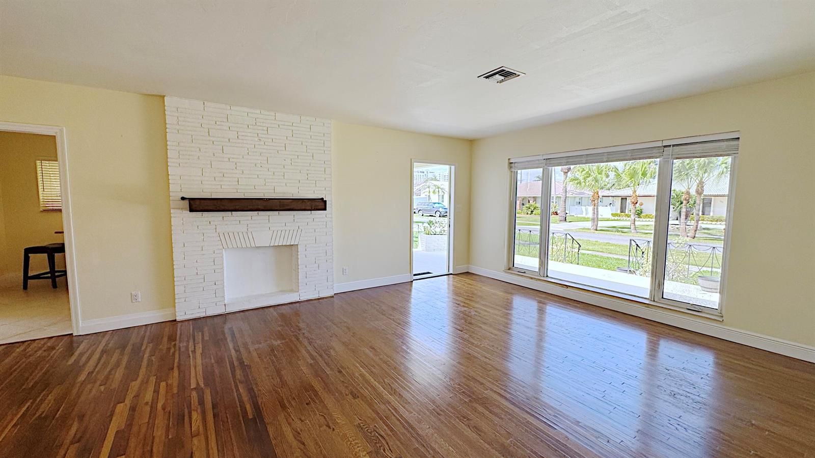 An empty living room features hardwood floors, a white brick fireplace, and large windows — 222 SE 2nd Terrace, Dania Beach FL 33004, DANIA BEACH, FL