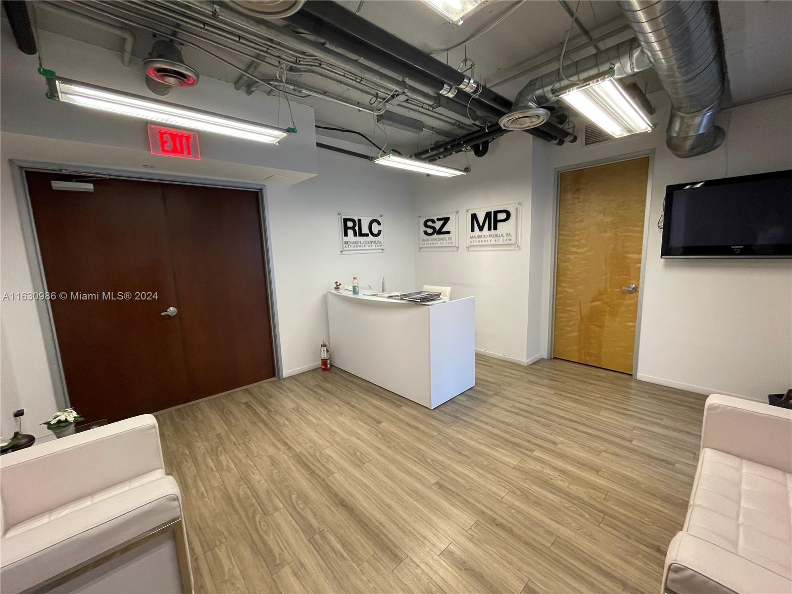A reception area features a white desk, wood-look flooring, exposed ceiling pipes, and three signs — 14 NE 1st Ave # 300A, Miami FL 33132, MIAMI, FL