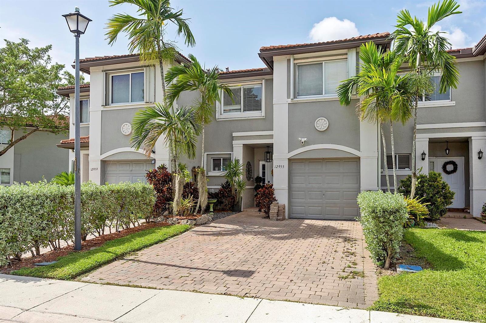 A two-story townhouse exterior features a brick driveway, palm trees, and a gray garage door — 12955 SW 28TH Court, Miramar FL 33027, MIRAMAR, FL