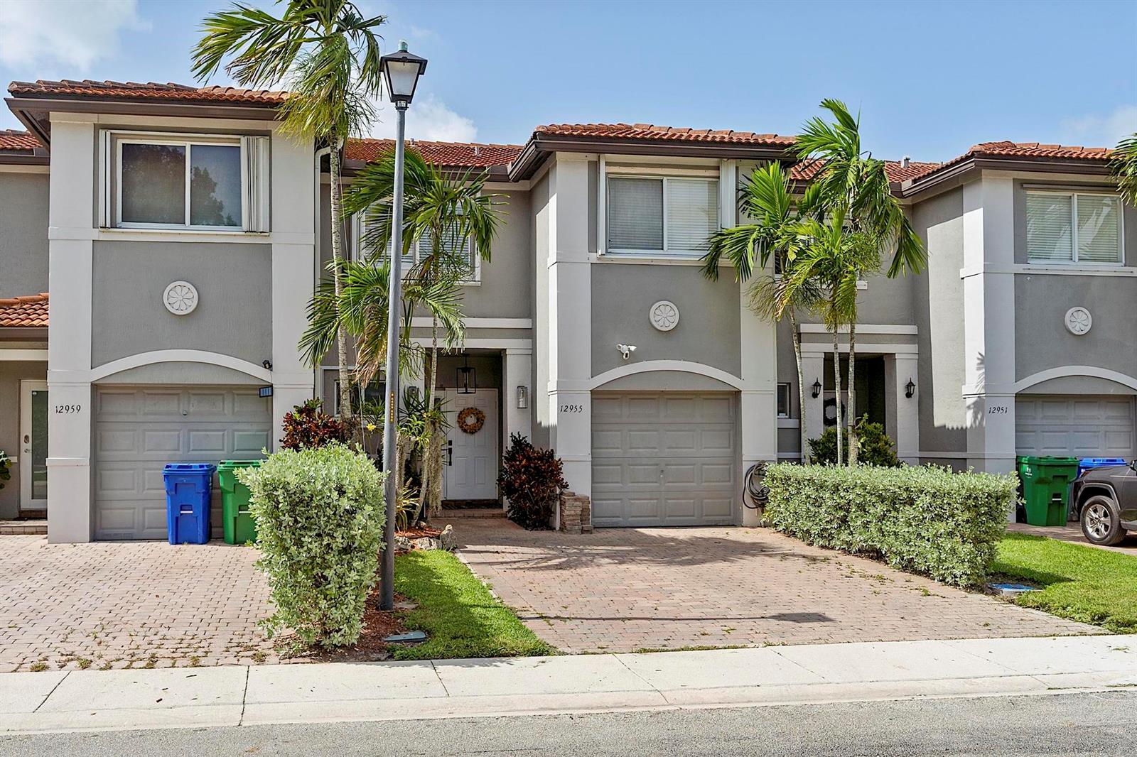 Three attached townhomes with gray stucco facades, tiled roofs, and paved driveways under a blue sky — 12955 SW 28TH Court, Miramar FL 33027, MIRAMAR, FL