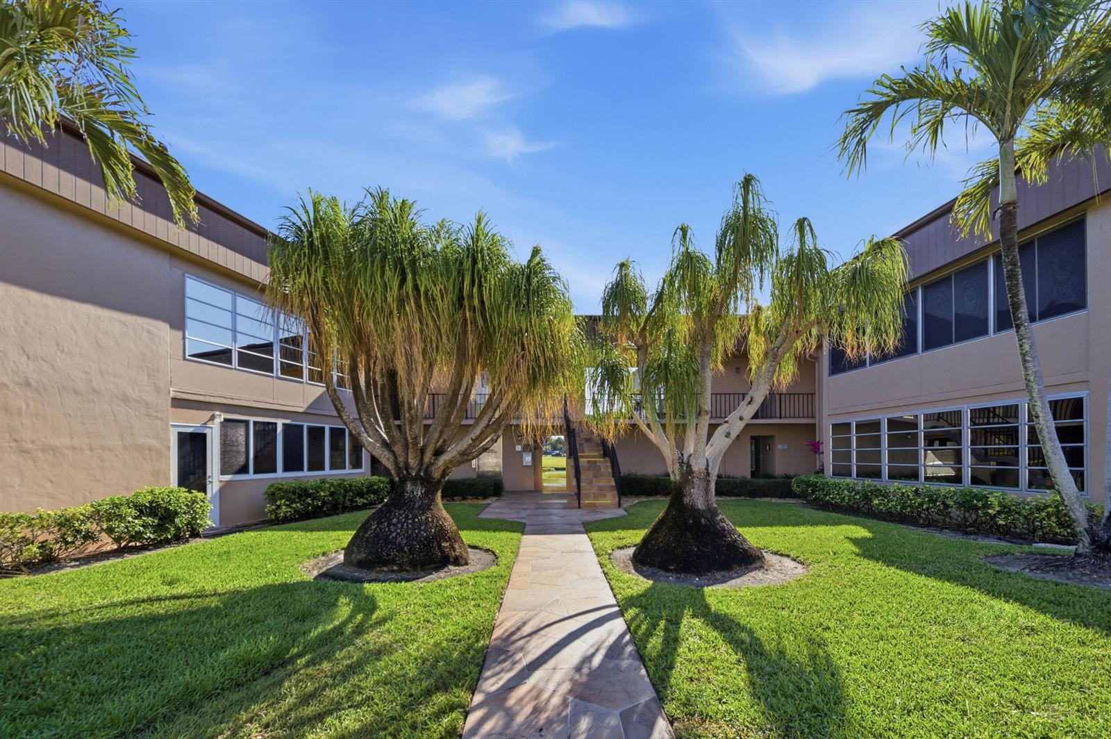 A stone path leads between two large palm trees toward an outdoor staircase between buildings — 129 Piedmont C, Delray Beach FL 33484, DELRAY BEACH, FL