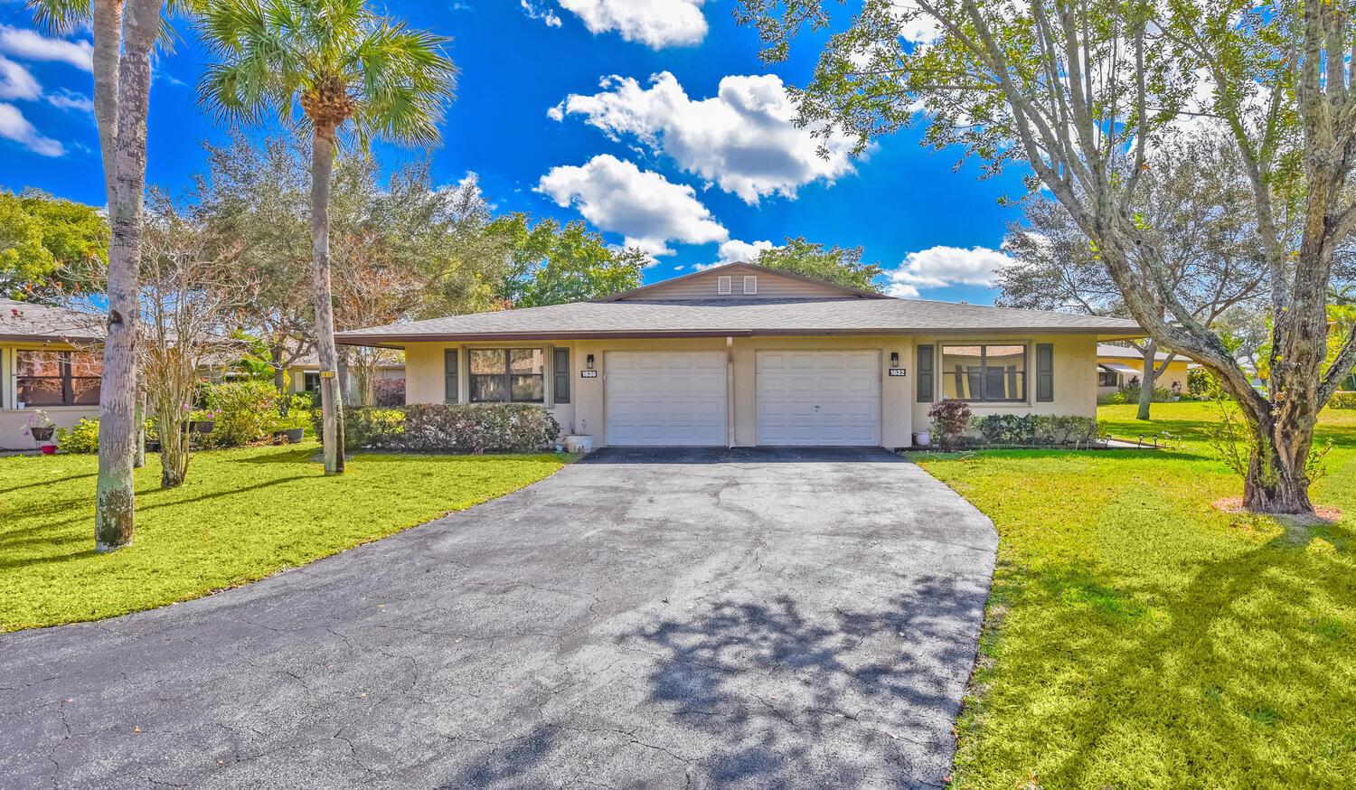 A single-story duplex with two garage doors, a shared driveway, and surrounding trees under blue skies — 1630 Palmland Drive, Boynton Beach FL 33436, BOYNTON BEACH, FL