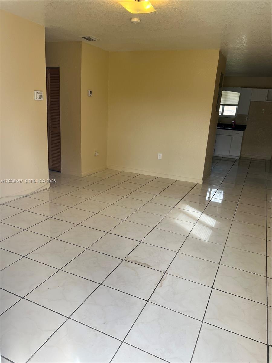 An empty room features light-colored tile flooring, beige walls, and a view into a kitchen — 789 NW 55th Ter # B, Miami FL 33127, MIAMI, FL