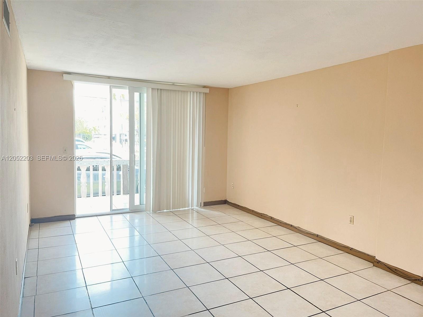 An empty room features light-colored tile flooring, beige walls, and a sliding glass door — 1475 NE 125th Ter # 112A, North Miami FL 33161, NORTH MIAMI, FL