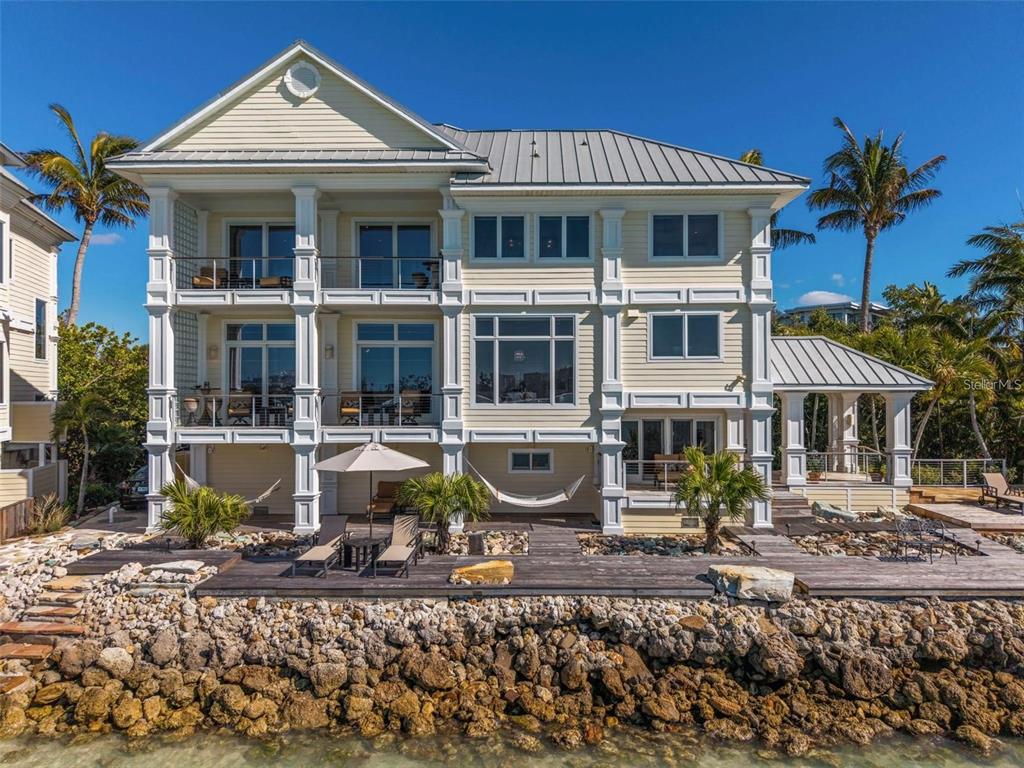 A three-story yellow house with a metal roof sits behind a rocky shoreline — 3750 BAYOU LOUISE LANE, SARASOTA, FL