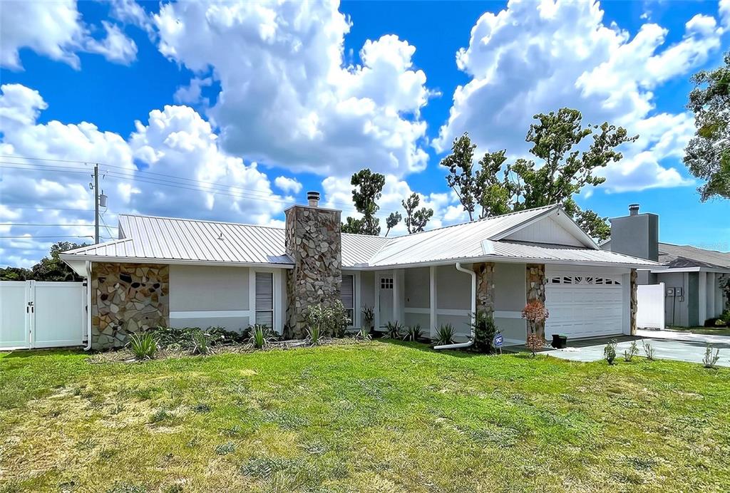 A single-story house with a stone chimney, metal roof, and attached garage under a blue sky — 5520 FULMAR DRIVE, TAMPA, FL