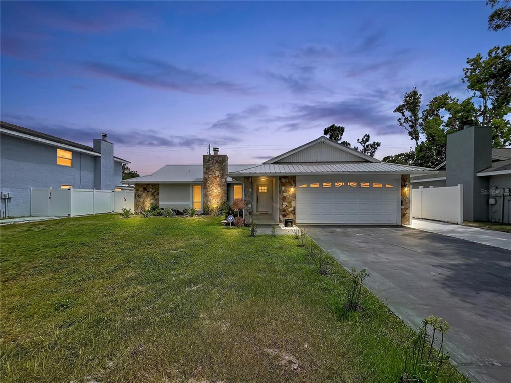 A single-story house with a stone chimney, white garage door, and metal roof at dusk — 5520 FULMAR DRIVE, TAMPA, FL