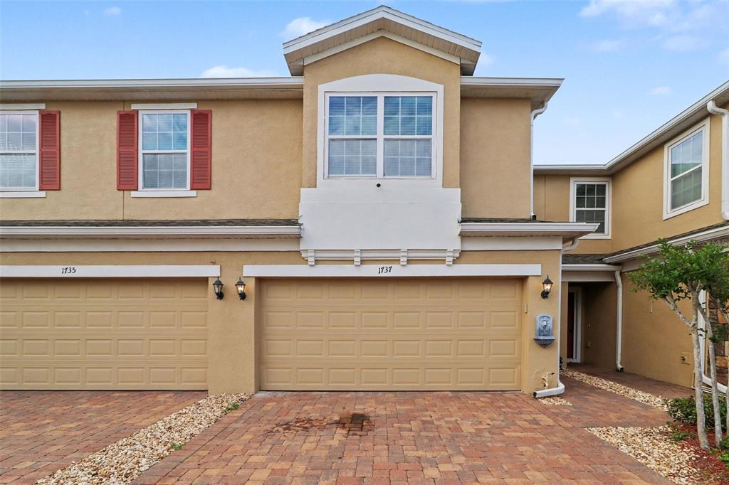 A two-story townhouse exterior features a two-car garage, tan stucco siding, and brick pavers — 1737 KATO WAY, KISSIMMEE, FL