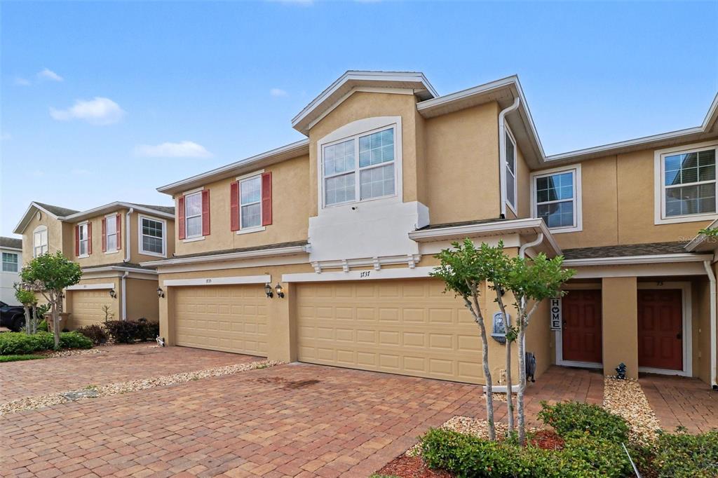 A two-story townhouse exterior features tan stucco walls, a brick paver driveway, and garage doors — 1737 KATO WAY, KISSIMMEE, FL