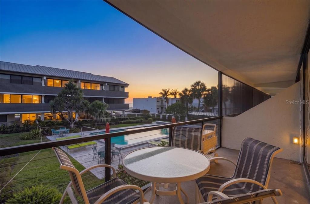 A balcony with a table and chairs overlooks a swimming pool at sunset — 6804 GULF DRIVE UNIT 6804, HOLMES BEACH, FL