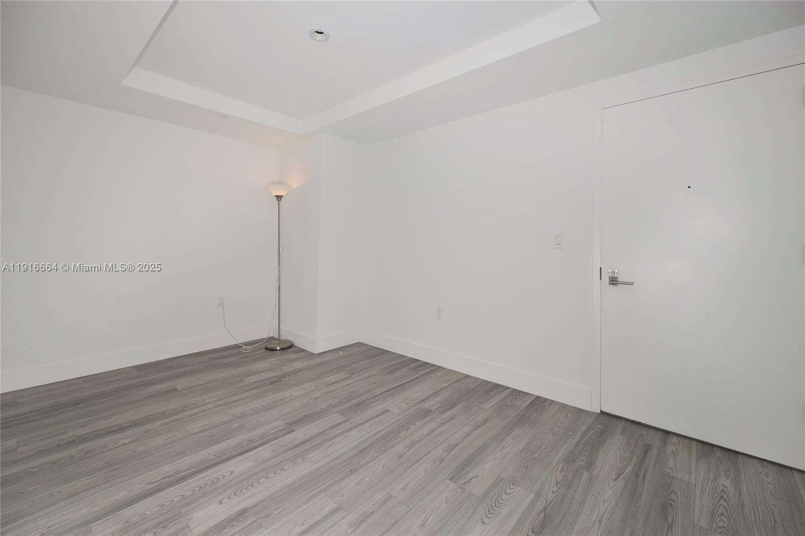 An empty room features white walls, gray wood-look flooring, a floor lamp, and a door — 45 SW 9th St # 1504, Miami FL 33130, MIAMI, FL