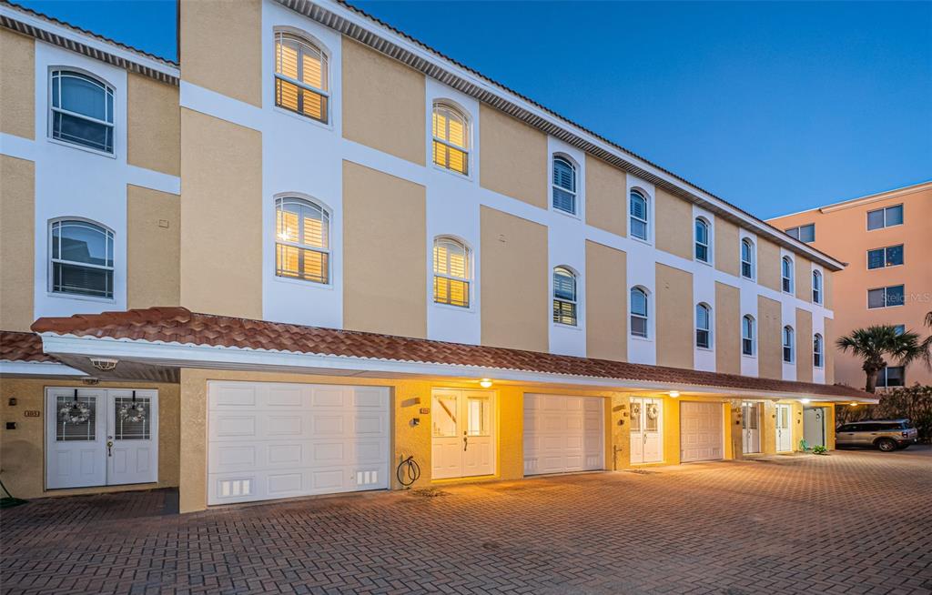 A multi-story tan building with arched windows, white garage doors, and a brick-paved driveway — 11595 GULF BOULEVARD UNIT 6, TREASURE ISLAND, FL