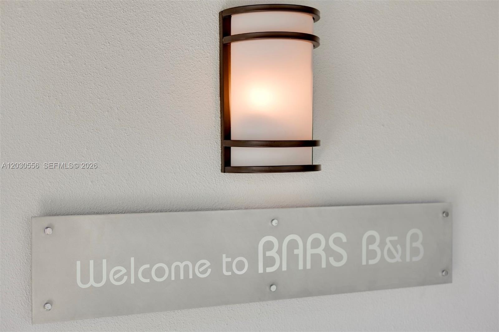 A wall-mounted light fixture hangs above a metal sign that reads Welcome to BARS B&B — 711 Lenox Ave, Miami Beach FL 33139, MIAMI BEACH, FL